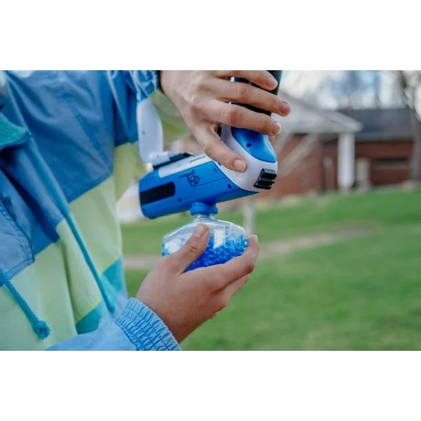 A person in a blue and green jacket is loading a blue and white gel blaster toy gun with a clear container filled with blue gel beads. The scene takes place outdoors on a grassy area with a house in the background.